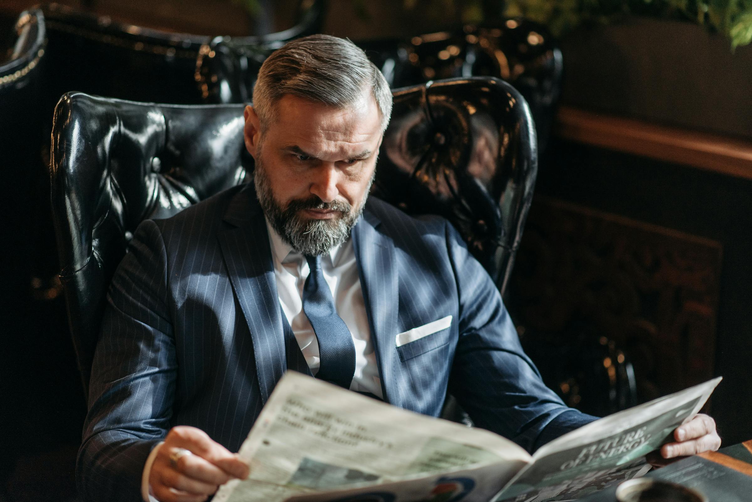 A senior executive in a pinstripe three-piece suit reads a newspaper in a tufted leather chair — the intended reader of the weekly briefing